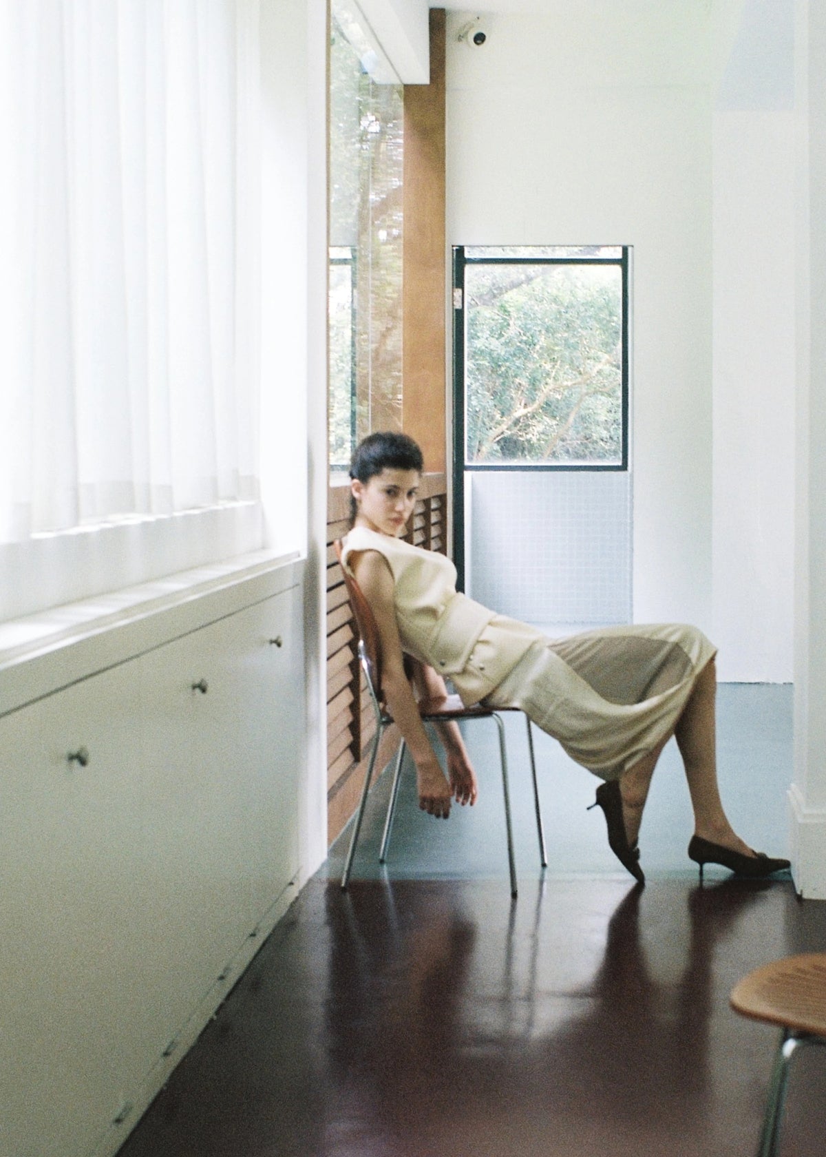 Model seated beside tall windows in a pale dress.
